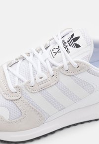 Bílá sportovní obuv z prodyšné síťoviny a semiše s třemi bílými pruhy, polstrovaným límcem a černým logem na jazyku.