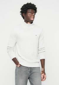 Lys grå turtleneck genser laget av strikket stoff, med ribbede mansjetter og liv. Brukes med grå denim jeans. Lite logodetalj på brystet.