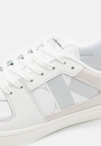 Sneaker bianco in pelle con accenti grigi. Presenta un prominente logo "CK" grigio, un pannello in suede testurizzato e lacci bianchi. Design liscio e arrotondato.