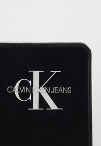 Černý kožený povrch s bílými iniciály "CK" a stříbrným logem "Calvin Klein Jeans" vyraženým uprostřed.