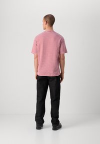 Rød og hvit stripete t-skjorte med korte ermer, rund hals, iført mørke denimbukser og svarte sko, sett fra baksiden.