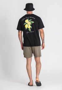 Čierne tričko s grafickým motívom citrónu na chrbte, v kombinácii s béžovými šortkami a čiernymi sandálmi. Minimalistický design s krátkymi rukávmi.