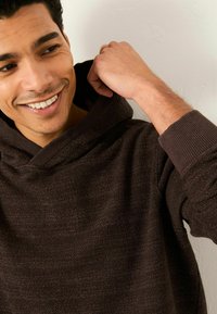 Brauner Kapuzenpullover mit strukturierter Oberfläche, weiter Kapuze, gerippten Bündchen und lockerem Schnitt. Der Stoff wirkt weich mit dezenten Farbnuancen.