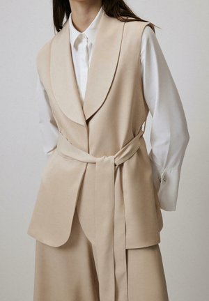 Vrouw draagt een beige mouwloze, ceintuurvest over een wit knoopjesshirt met lange mouwen tegen een effen achtergrond.