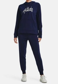 Marineblaues Hoodie mit Fronttasche und Kordelzugkapuze, mit weißem gedrucktem Logo; kombiniert mit marineblauen Jogginghosen und weißen Sneakers.