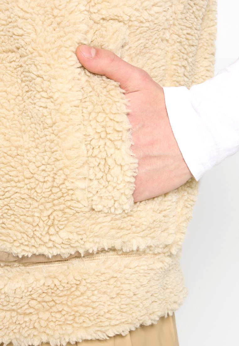 Gilet di pile beige con una superficie morbida e strutturata e una tasca laterale per le mani. Presenta una vestibilità comoda e cuciture lungo l'orlo inferiore.