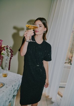 Une femme en robe noire lève un verre de boisson dorée près d’une table décorée et d’une fenêtre à rideaux.