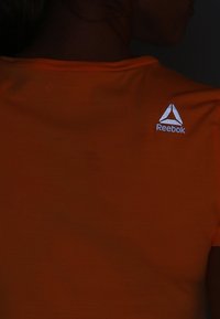 Orange short-sleeve shirt made of a smooth fabric, featuring a reflective white Reebok logo on the back, with a crew neckline.