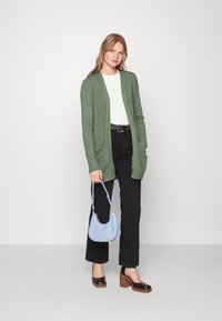 Groene gebreide cardigan met open voorkant, twee zakken, gedragen over een witte top, gecombineerd met zwarte high-waisted broek en bruine block-heeled schoenen.