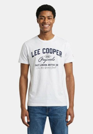 Joven sonriente que lleva una camiseta blanca de Lee Cooper con texto azul y vaqueros azules, de pie frente a un fondo blanco liso.