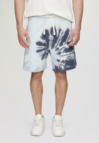 Shorts tie-dye bleu clair avec des motifs en tourbillon bleu marine, taille élastique, cordon blanc et ourlets roulés, associés à des baskets blanches.