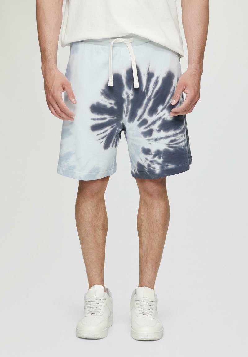 Lichtblauwe tie-dye shorts met marineblauwe spiraalpatronen, elastische tailleband, witte trekkoord en opgerolde zoom, gecombineerd met witte sneakers.