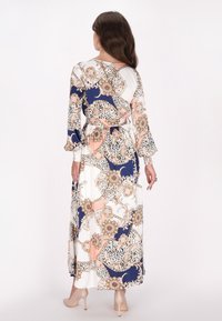Kvindelig med langt brunt hår iført en lang, mønstret kjole med blomster- og abstrakte designs i marineblå, ferskenfarvet og hvid, parret med beige hæle.
