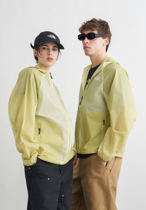 Deux personnes portant des vestes translucides jaune pâle, l'une avec une casquette noire et un pantalon noir, l'autre avec des lunettes de soleil et un pantalon beige, posant devant un fond blanc.