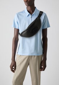 Schwarze Leder-Schultertasche mit geschwungener Form und geprägtem Logo, diagonal über ein hellblaues Hemd mit Kragen und beigen Hosen getragen.