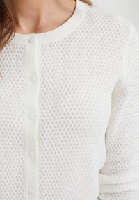 Bílý pletený cardigan s strukturovaným vzorem připomínajícím včelí plástev, kulatým výstřihem a čtyřmi malými knoflíky vpředu. Měkká tkanina s mírným elastickým efektem.