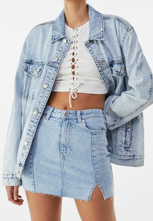 Hellblaue Jeansjacke in oversize Passform, mit Metallknöpfen und zwei Vordertaschen. Kombiniert mit einem weißen, gerippten Croptop und einem passenden Rock.