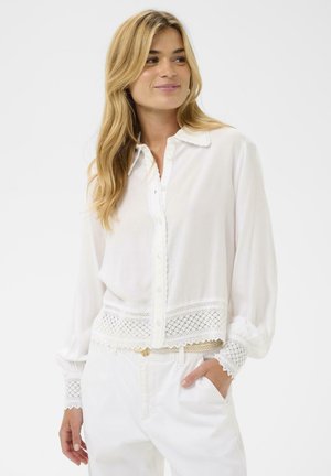 Blouse blanche avec fermeture à boutons, bordure en dentelle au bas et aux manches, manches longues et col à revers. Texture lisse, tissu léger.