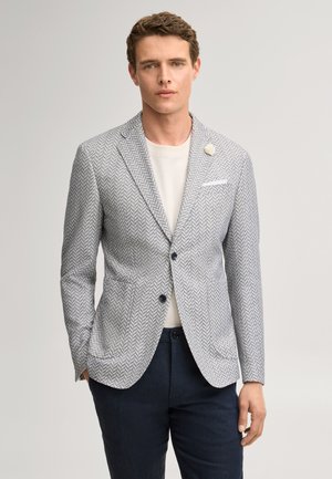 Man wearing gray herringbone blazer with white flower pin, white shirt, and dark trousers, standing against plain light background.