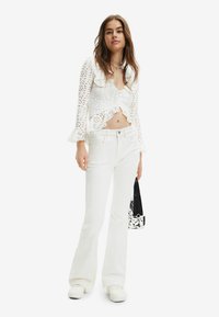 Top corto bianco con volant e design a traforo, abbinato a jeans bianchi a palazzo e sneaker bianche, mentre tiene una borsa a motivo bianco e nero.