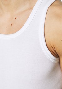 Hvid bomulds tanktop med ribbet tekstur, der har en rund halsudskæring og brede armåbninger, der viser hud og fregner nedenunder.