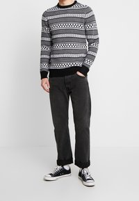 Sweter dziergany na drutach w czarno-białe geometryczne wzory, z okrągłym dekoltem i ściągaczami na mankietach, zestawiony z ciemnymi jeansami i czarnymi trampkami.