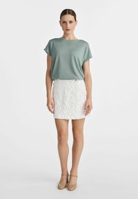 Jasnozielony top z krótkim rękawem zestawiony z białą mini spódniczką o teksturowanym wzorze z florystycznymi motywami. Modelka ma na sobie beżowe buty na słupku.