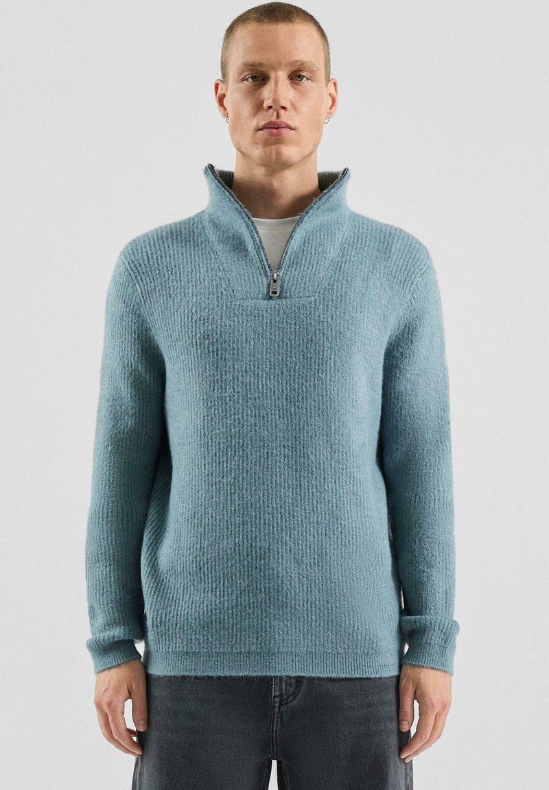 Jasnoniebieski, teksturowany sweter dziany z wysokim kołnierzem i suwakiem do ćwiartki długości. Mankiety i dół z prążkowanym wzorem dodają detalu. Noszony z jasnoszarą koszulą pod spodem.