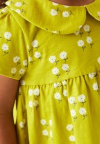 Bambina che indossa un vestito verde con maniche a sbuffo corte e motivo floreale di margherite bianche con piccole foglie.