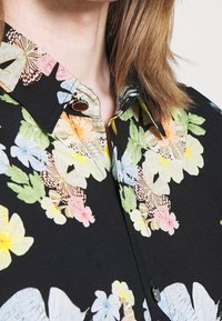Blusa floreale in tessuto nero con fiori colorati e farfalle, colletto classico con dettaglio dei bottoni e texture liscia.