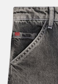Juodi džinsai iš denim su tekstūruotu paviršiumi, su kontrastingais baltais siūlais ir mažo raudono "HUGO" ženkleliu ant monetų kišenės.