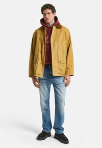 Geel katoenen jack met opgestikte zakken, bordeauxrode hoodie eronder, blauwe jeans en zwarte schoenen. Model staat tegen een witte achtergrond.
