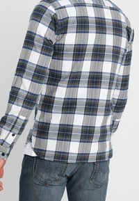 Camisa de cuadros para hombre fabricada en algodón. Presenta un patrón de rayas en azul, gris y blanco, con mangas largas y puños abotonados. Corte recto en el dobladillo.