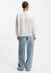 Weiße Bluse mit langen Ärmeln, gerafftem Rückendetail und Knopfverschluss. Kombiniert mit hellblauen, weit geschnittenen Jeans mit ausgefransten Säumen.