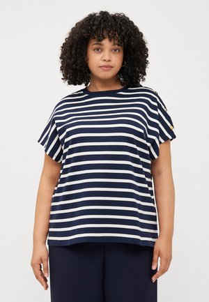 Une personne aux cheveux bouclés porte un t-shirt à rayures bleu marine et blanc avec des manches courtes et des détails en boutons, se tenant devant un fond uni.