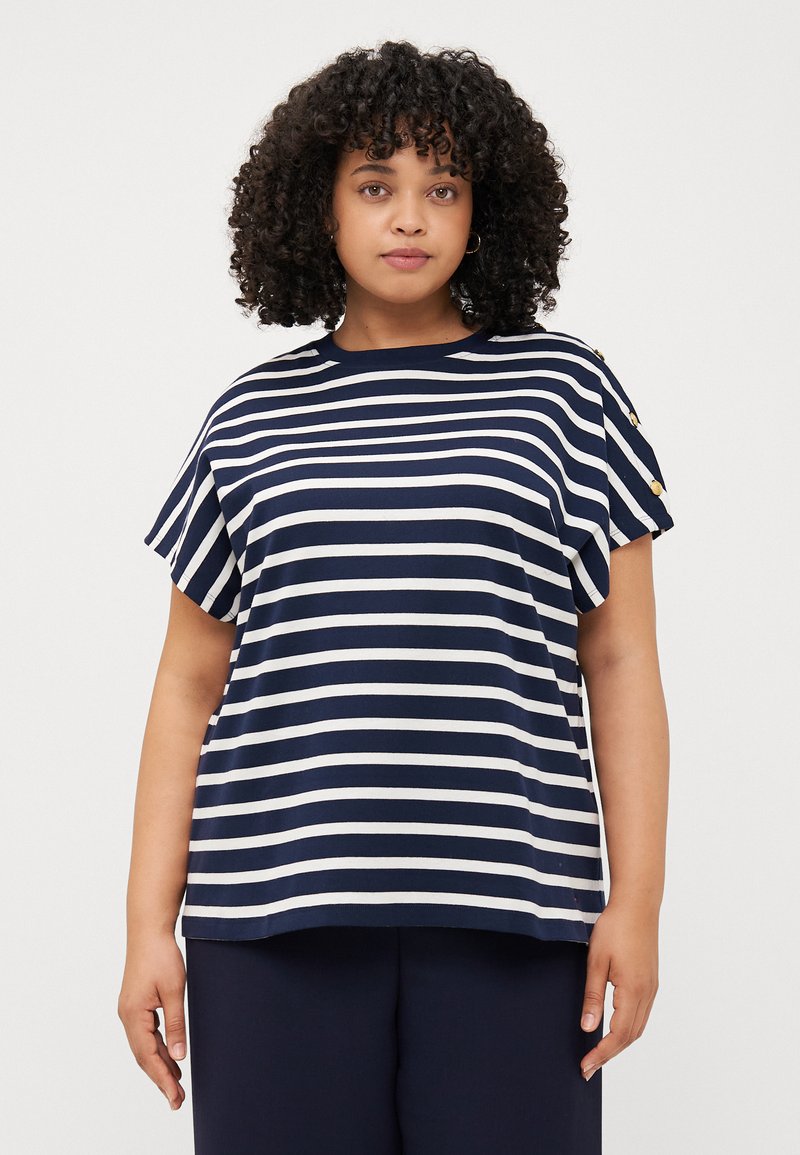 En person med krøllet hår har en navy og hvid stribet t-shirt med korte ærmer og knapdetaljer, stående mod en ensfarvet baggrund.