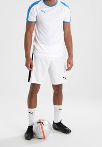 Man i vit och blå Puma fotbollsklädsel står med ena foten på en silverfärgad Puma fotboll mot en vit bakgrund.