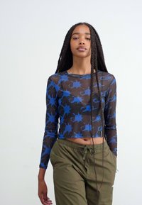 Dolga majica s kratkimi rokavi iz prosojnega materiala, s modrim cvetnim vzorcem na črni podlagi. V kombinaciji z olivno zelenimi hlačami.