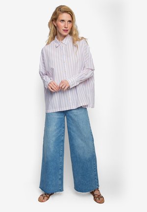 Overhemd met knopen en lichtpaarse en oranje strepen, gecombineerd met brede denim jeans. Model staat met handen gevouwen en draagt bruine sandalen.