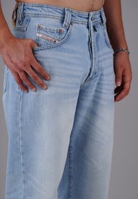 Hombre que lleva puestos unos pantalones de mezclilla azul claro con la mano descansando cerca del bolsillo delantero y una pulsera de plata en la muñeca.