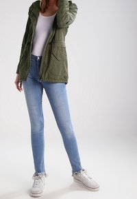 Olivově zelená bunda s kapucí, bílý top bez rukávů, světle modré úzké džíny a bílé tenisky s třásněmi na lemu.