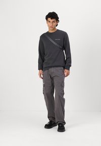 Sudadera gris oscuro con logo, combinada con pantalones cargo a juego con bolsillos laterales y zapatos negros. El hombre se encuentra de pie con confianza, con las manos relajadas.