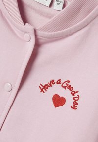 Top in tessuto rosa chiaro con scollatura rotonda, bottoni a pressione e testo ricamato rosso "Buona Giornata" con un particolare a forma di cuore.