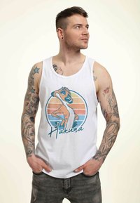 Hvid tanktop med et blåt og orange grafik, der viser en tegneseriefigur og "Hakuna" tekst. Modellen bærer grå jeans og viser tatoverede arme.