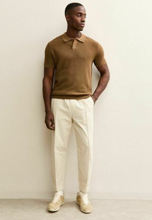 Polo marrone in maglia con maniche corte, abbinata a pantaloni color crema e sneakers beige, in piedi davanti a uno sfondo neutro.