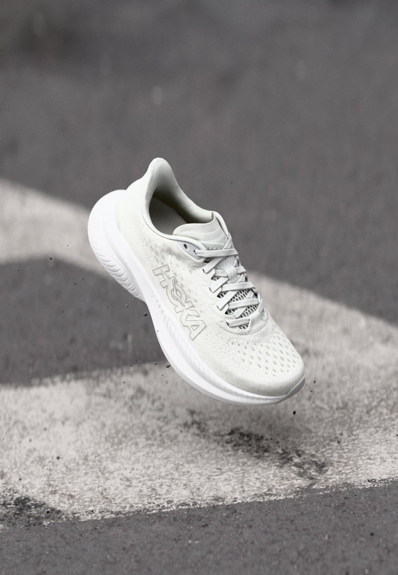 Baskets de course légères en gris clair avec une tige en maille, semelle blanche et texture perforée. Présente des lacets et le logo "HOKA" sur le côté.