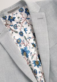 Blazer gris clair avec une doublure en satin à motifs floraux comprenant de grandes fleurs bleues et des feuilles vertes sur fond blanc.