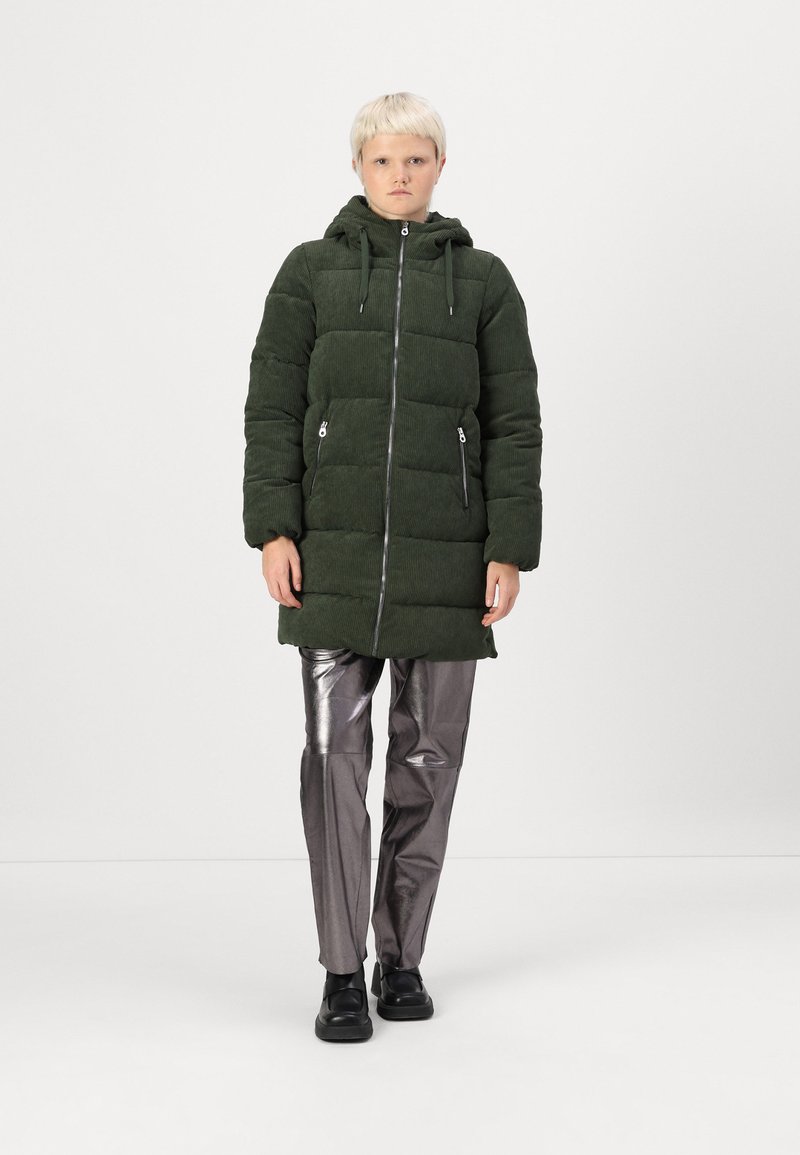 Chaqueta de plumón de canalé verde con capucha, cierre de cremallera y dos bolsillos frontales. Combinada con pantalones plateados brillantes y zapatos negros.