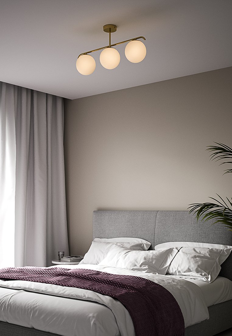 Lampadario a tre globi con finitura in ottone; paralumi bianchi rotondi in vetro; pende sopra un letto imbottito grigio con una coperta viola.