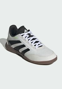 Witte en zwarte lage Adidas sportschoen met witte veters, zwart logo met drie strepen en een bruine zool op een lichte achtergrond.